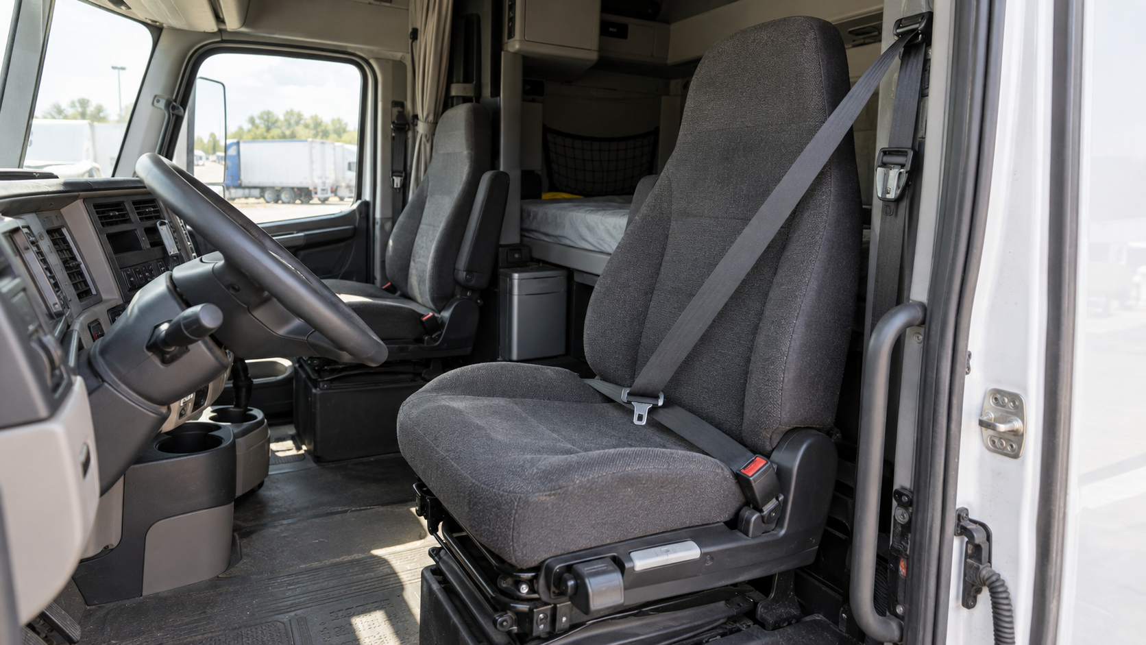 Documentary view through the open passenger-side door of a realistic unbranded American Class 8 sleeper cab, showing a lightly worn front seat with a correctly routed three-point belt, the driver controls and sleeper compartment in neutral daylight