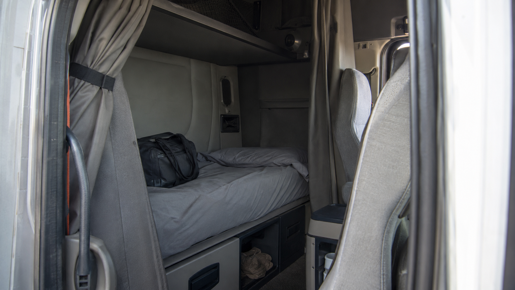 Documentary view through the open doorway of a realistic American Class 8 sleeper tractor, showing a clean but lightly worn lower bunk with plain gray bedding, privacy curtains, practical storage and an unbranded duffel in neutral morning light